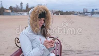 穿着白色羽绒服的女人坐在海滩上的长凳上看着手机。
